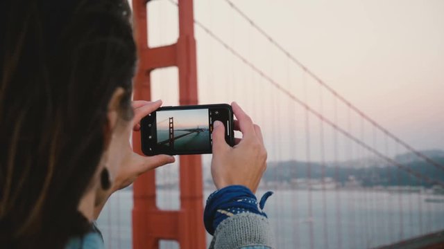 Back view close-up young woman takes smartphone photo of amazing sunset Golden Gate Bridge in San Francisco USA.
