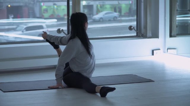 Young woman stratching legs on mat in the gym with large floor-to-ceiling windows. Modern urban cityscape, cars behind the glass. Girl exercising healthy lifestyle in yoga studio. Camera moves closer