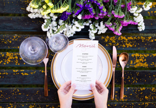 Hands Holding a Menu Mockup on Table with Tableware and Flowers