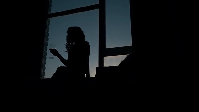 black silhouette of slim girl with curly hair holding wine glass on windowsill in dark room against evening sky
