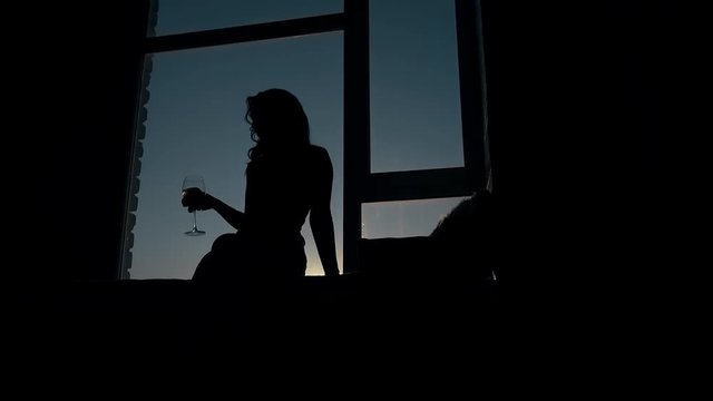 attractive silhouette of young graceful woman with glass of wine sitting on windowsill in dusk
