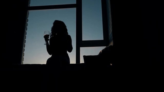 silhouette of happy dancing lady with long curly hair holding wineglass sitting on windowsill in dark room