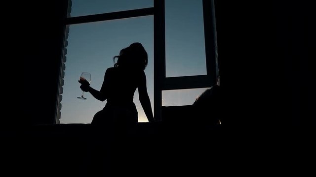 silhouette of graceful girl with wine sitting and dancing by window in darkness against deep blue evening sky