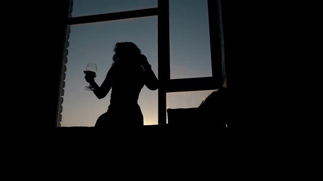 silhouette of curly haired lady dancing and holding wineglass in dark room against sunset light behind window
