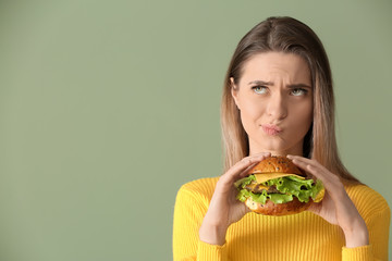 Thoughtful young woman with tasty burger on color background