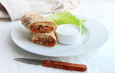 shawarma with chicken, vegetables and herbs on a white plate next to the sauce and knife on a light background.