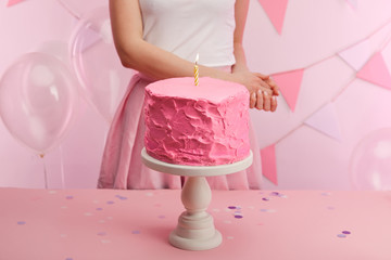 cropped view of woman standing near sweet and tasty pink birthday cake with burning candle on cake stand