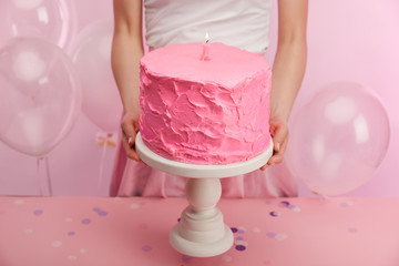 cropped view of woman holding cake stand with pink sweet birthday cake and burning number one candle near air balloons