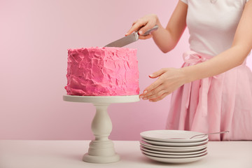 Fototapeta na wymiar cropped view of woman holding knife near pink birthday cake on cake stand near saucers on pink