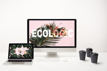 computer with ecologic lettering on monitor and laptop with eco lettering on screen on desk with paper cups on white background