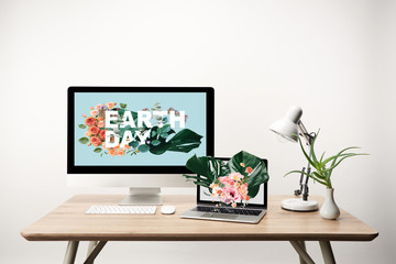 computer with earth day illustration on monitor and laptop with monstera leaves and flowers illustration on screen on wooden table