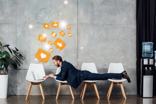 businessman lying on chairs and using laptop in waiting hall with social media icons on wall