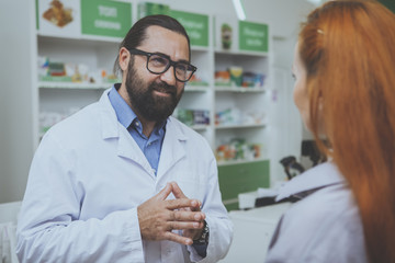 Fototapeta premium Cheerful male pharmacist helping female customer buying medications at the drugstore. Bearded chemist working at pharmacy, giving medical advise to the patient. Communication, medical care concept