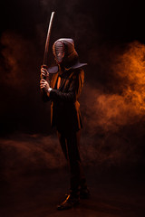 Full length view of kendo fighter in formal wear and helmet holding bamboo sword on dark