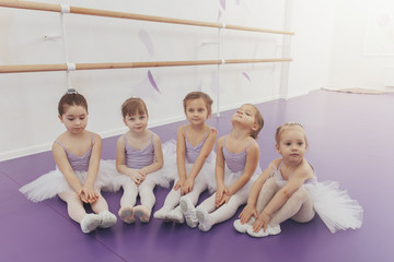 Fototapeta na wymiar Cute little girls sitting on the floor, resting after ballet lesson at dance studio. Adorable little ballerinas wearing tutus relaxing together after exercising. Health, lifestyle, innocence concept