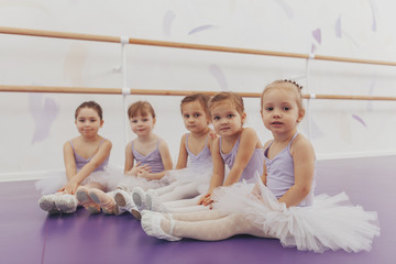 Lovely little girls looking to the camera, sitting on the floor at ballet school. Adorable little ballerinas rest after exercising. Group of little girls dancers wearing leotards relaxing after ballet