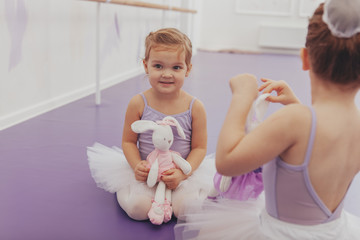 Cute happy little girl in leotard and tutu holding her toy, sitting with her best friend at ballet...