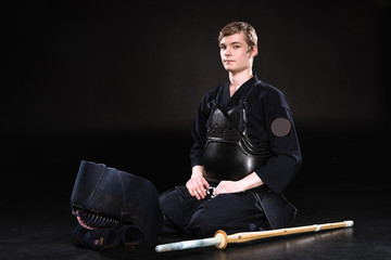 Young man in kendo armor with helmet and bamboo sword sitting on black