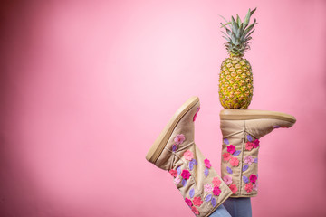 Cute female legs wearing boots and holding pineapple over pink background
