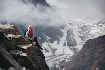 smiling blonde with a backpack sitting on a rock. the concept of traveling in mountains