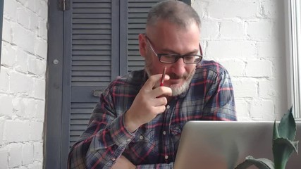 Gray-haired bearded male accountant working at home behind a laptop, making a report against the backdrop of a minimalist interior, takes a call from a customer on Skype, drinks tea or coffee, smiles. - Powered by Adobe