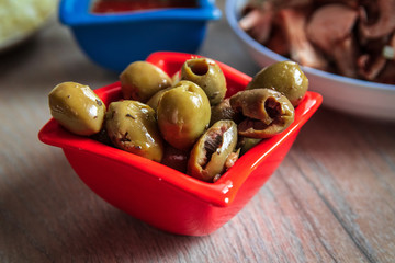 Green olives with spices for pizza in a red bowl and other pizza ingredients in the background