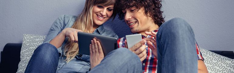 Portrait of young couple in love looking electronic tablet and laughing while resting over a bed. Leisure time at home concept.