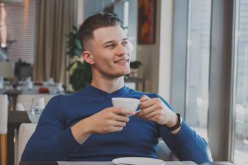 Attractive man smiling, looking in the window, relaxing at the cafe holding cup of delicious coffee. Happy man having breakfast at the restaurant looking away dreamily. Lifestyle, happiness concept