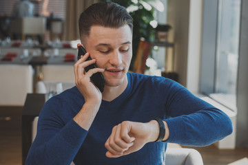 Handsome businessman talking on his phone, looking at the watch while waiting for a business meeting at the restaurant. Attractive man calling someone, checking time on his watch. Time management 