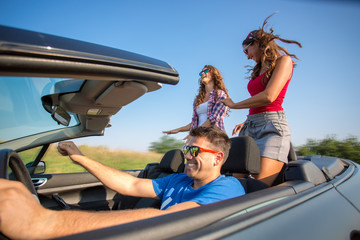 Friends driving in a convertible car while women are dancing in the back