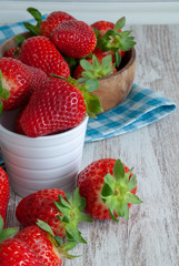 Vertical close-up of strawberries in white and wooden bowls. Over blue checkered kitchen rag and white wooden background