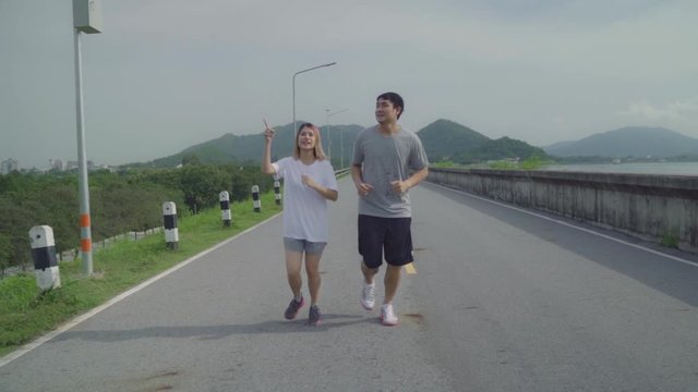 Slow motion - Asian runner couple running and jogging on street, Healthy young sporty sweet couple exercise near lake. Lifestyle fit and active couple exercise on street concept.