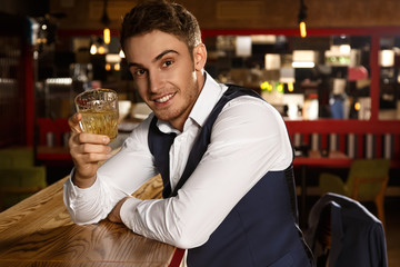 This one is to you. Handsome cheerful man smiling to the camera toasting with his glass of whiskey sitting at the bar 
