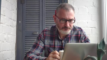 Gray-haired bearded male accountant working at home behind a laptop, making a report against the backdrop of a minimalist interior, drinking tea or coffee and reading information on the monitor. - Powered by Adobe