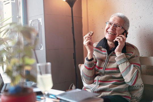 Senior Old Woman Enjoying A Fun Conversation Over Her Cell Phone, Eating A Chocolate Cookie In A Coffee Shop