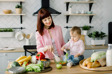A young and beautiful mother in a pink shirt is preparing a fresh vegetable salad at home in the kitchen, together with her little cute daughter sitting on the table. mother and daughter