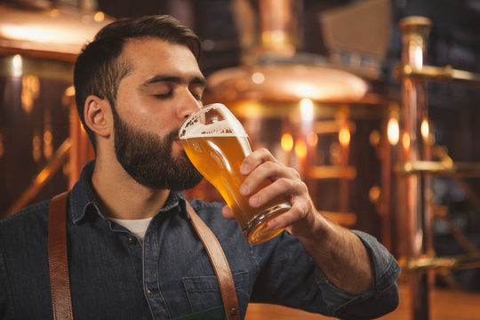Bearded male brewer sipping delicious ber from a glass, working at his production brewery. Professional brewer tasting fresh beer at his microbrewery, drinking with his eyes closed. Pleasure concept