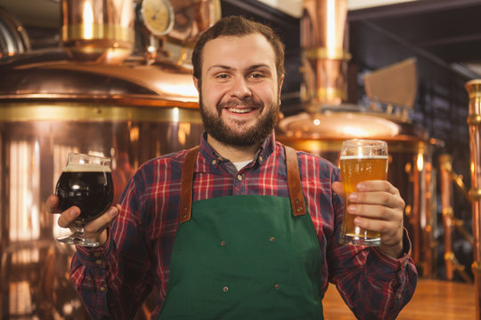 Cheerful friendly professional brewer in an apron smiling happily to the camera holding two glasses of beer, enjoying working at his microbrewery. Happy bartender serving two beers. Friendly service c