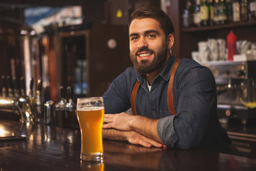 Handsome bearded bartender smiling joyfully to the camera, leaning on the bar counter, serving...