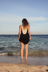 Fototapeta na wymiar A girl in a black dress on the coast of the sea, walking on the beach decided to wet her feet entering the water.