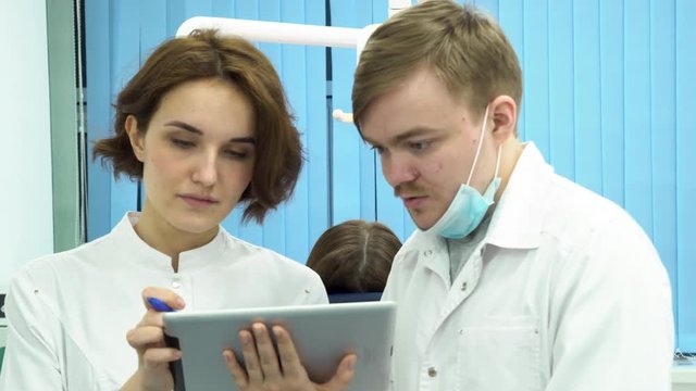 Serious doctors working together with tablet in the dental care cabinet with a patient lying behind. Dentists male and female discussing test reports that is shown on their tablet computer.