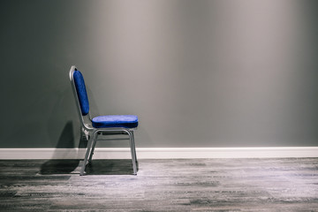 A lonely chair with silver legs in blue. It stands at the gray wall. At the bottom of a wide white baseboard and laminate. The shadow of the chair on the wall. Sun glare on the floor. Interior room