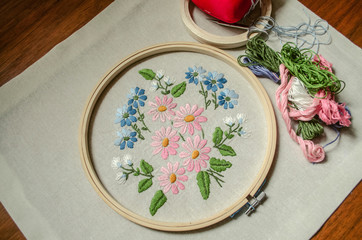 Embroidered bouquet of wild flowers on cotton fabric covered with wooden hoops, colored threads, scissors and a red pillow for needles