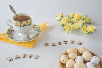 Easter breakfast in pastel colors with cup of tea,  chocolate eggs, yellow roses and wooden letters.  white background
