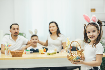 Pretty small girl with pigtails and bunny ears standing on the forefront with the yellow easter egg in her hand. Her father mother and brother sitting at the table, preparing for Easter.