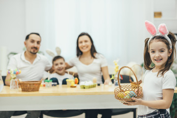 Pretty small girl with pigtails and bunny ears standing on the forefront with the basket of colored eggs in her hands. Her father mother and brother sitting at the table, preparing for Easter.