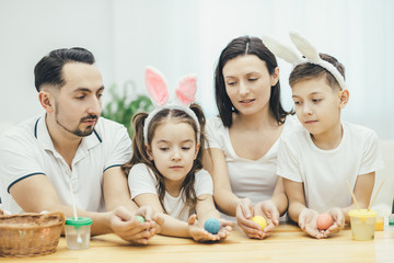 Inspired family in white t-shirts sitting together at the table, holding colored easter eggs in their hands, looking at them excitedly, choosing which egg is the best.