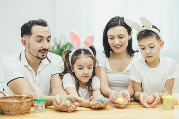 Inspired family in white t-shirts sitting together at the table, holding colored easter eggs in their hands, looking at them excitedly, choosing which egg is the best.