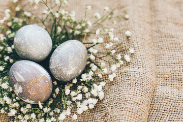 Stylish Easter eggs with spring flowers in floral nest on rustic fabric in sunny light on wood. Modern colorful eggs painted with natural dye in grey marble. Happy Easter, greeting card