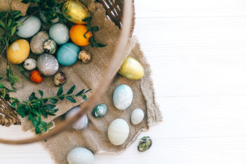Stylish Easter eggs and green buxus branches in rustic wicker basket on white wooden background. Flat lay. Modern gold easter candy and eggs painted with natural dye. Happy Easter.